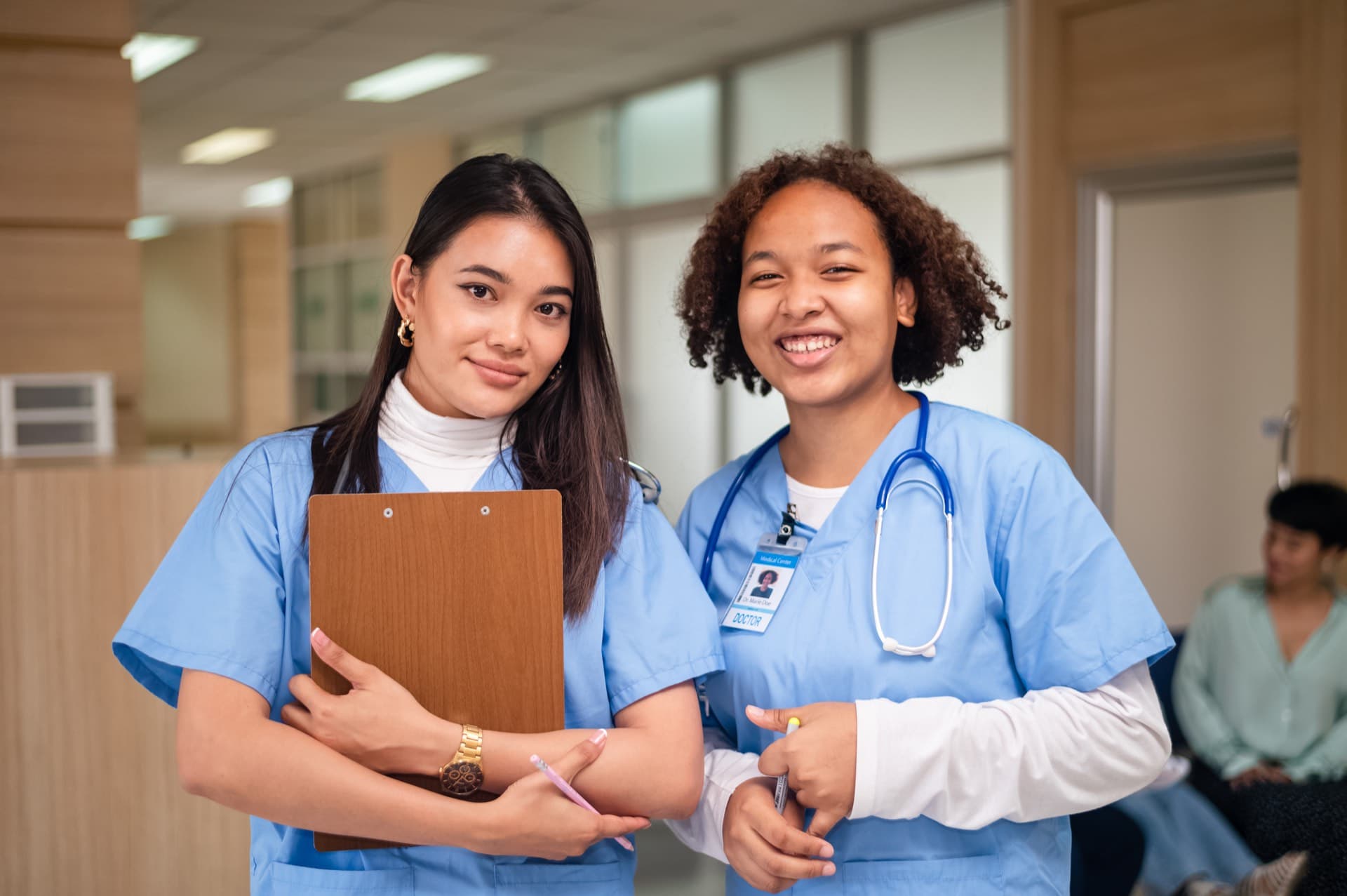 Two nursing students in scrubs at the hospital, smiling, one holding a clipboard.